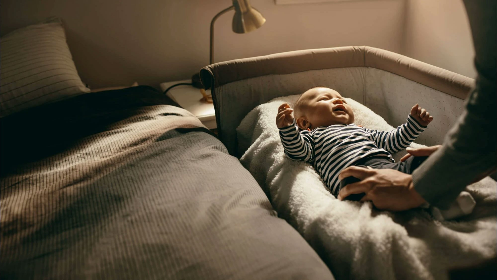 Baby in a striped body being put to bed in a bed side crib